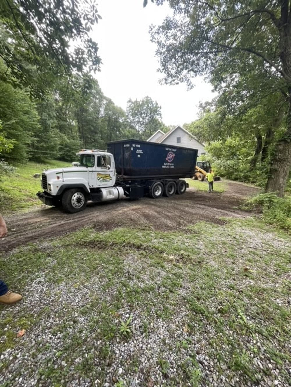 Large truck delivering a blue dumpster on a gravel driveway surrounded by trees.