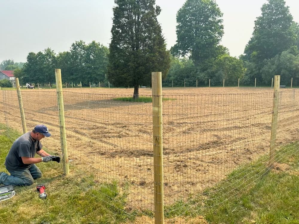 Man installing a fence around a field with trees in the background on a sunny day.