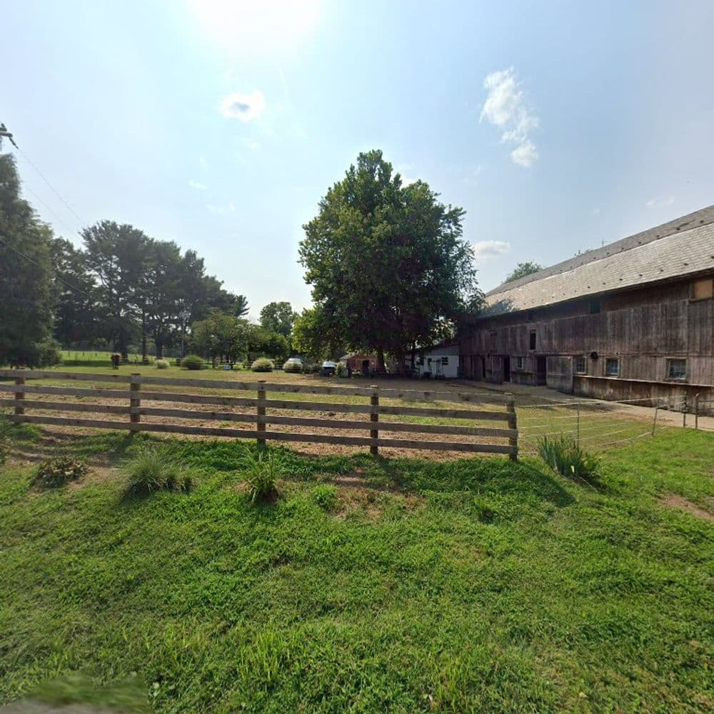 Rustic barn with wooden fence and green field under a bright sky, surrounded by trees.