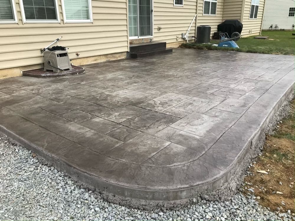 Stamped concrete patio installation behind a house with gravel surrounding the area.