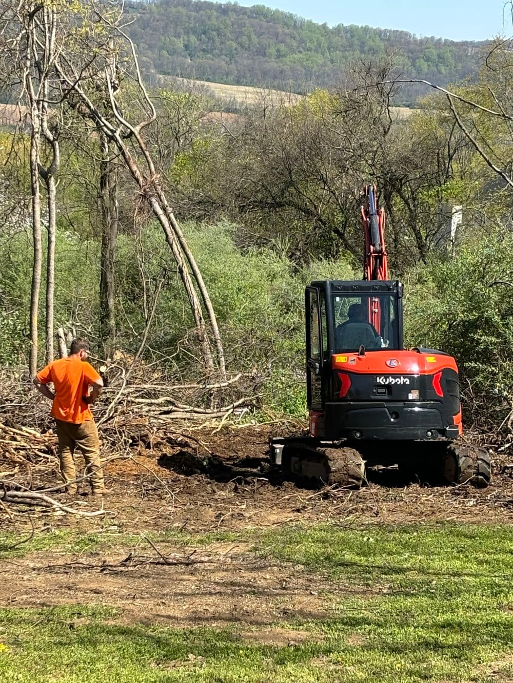 Excavator clearing land with operator in orange shirt among trees and brush.