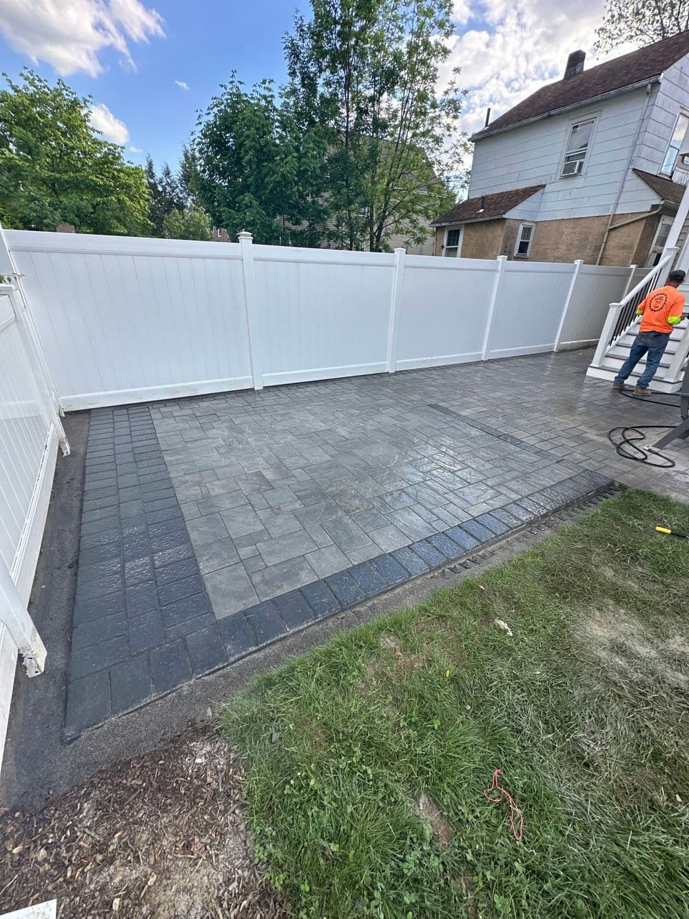 Paved patio area with contrasting pavers, fenced yard, and worker in safety vest.