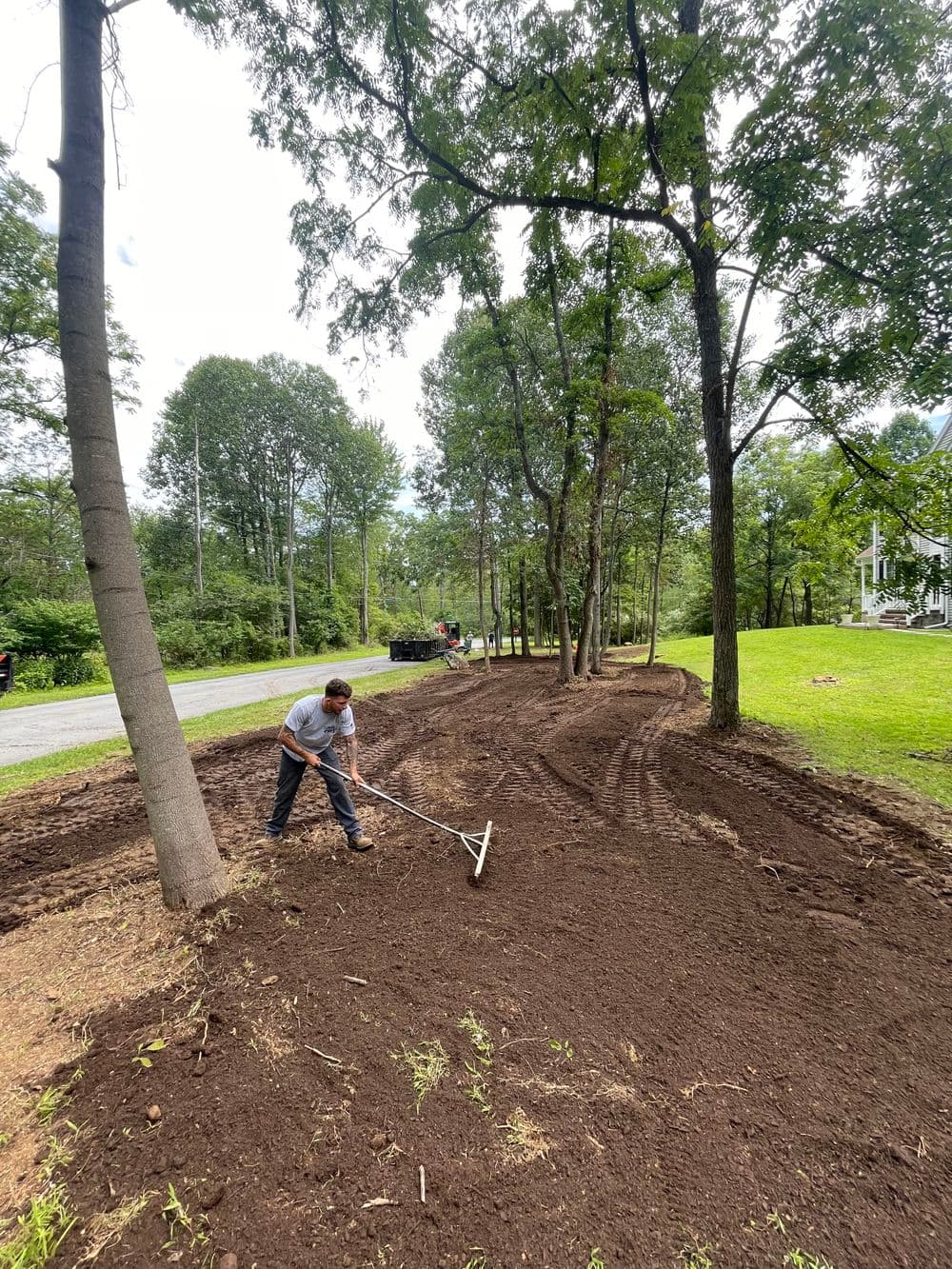Person raking freshly tilled soil in a landscaped yard surrounded by trees and a road.