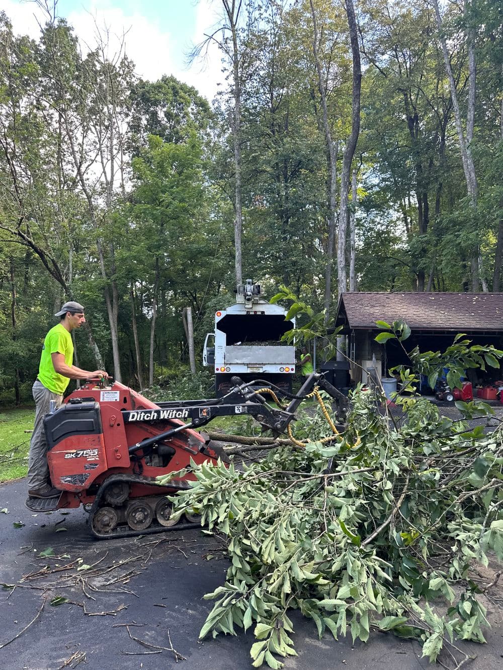 Man operating a Ditch Witch on a property, clearing fallen branches in a wooded area.