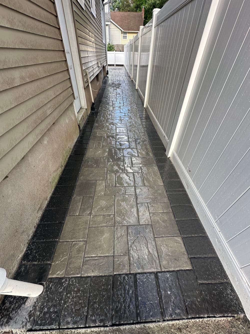 Stamped concrete walkway between two fences with a wet finish, featuring gray and black patterns.
