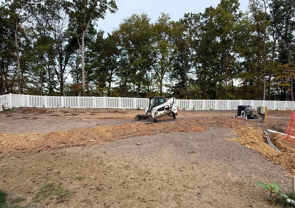 Construction site with a bobcat skid steer preparing land, surrounded by a white fence.