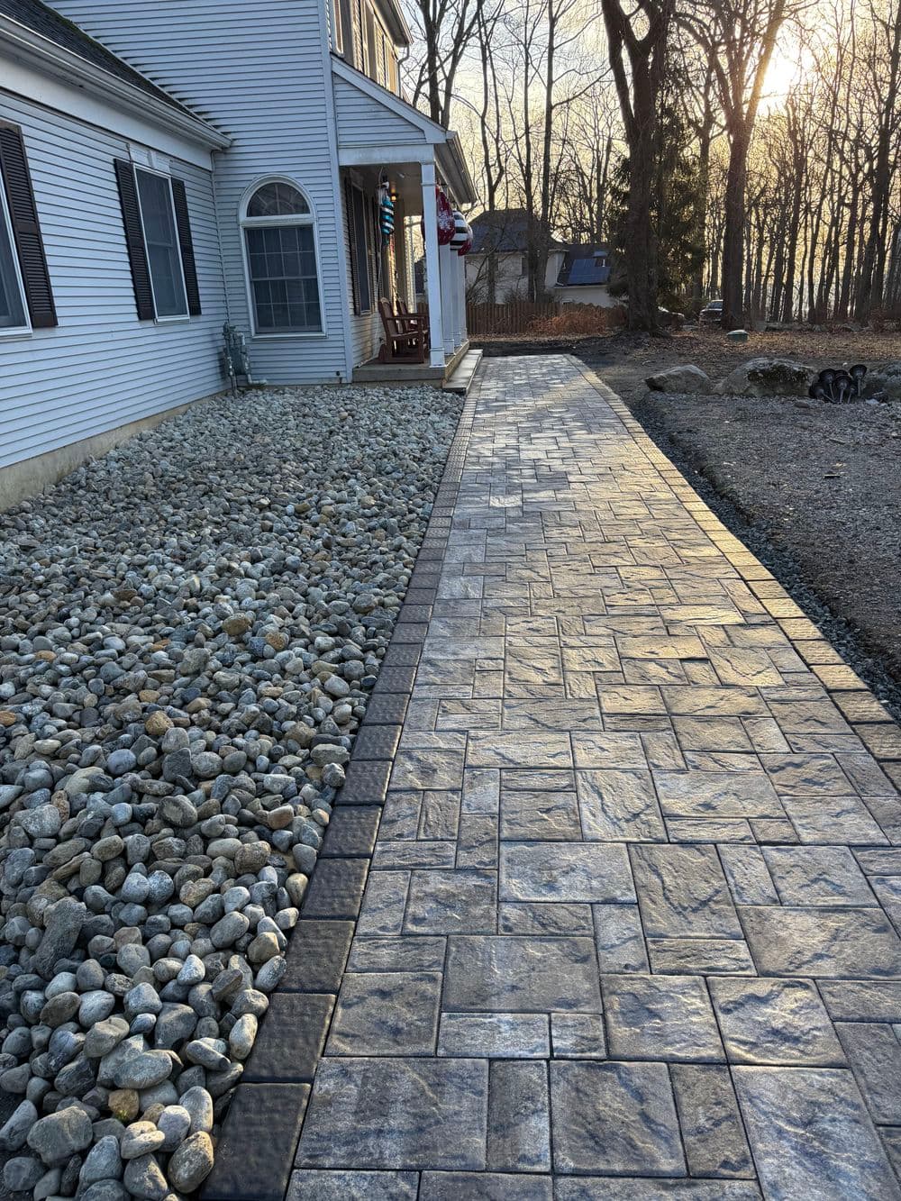 Textured stone walkway beside rocky landscaping leading to a house in a wooded area.